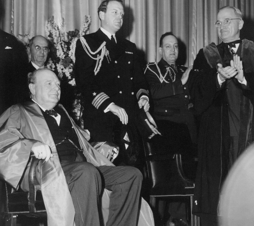 A black and white photograph showing Winston Churchill wearing academic robes seated on the right with dignitaries behind him and President Harry S. Truman standing on the left, also in academic robes looking at Churchill and applauding.