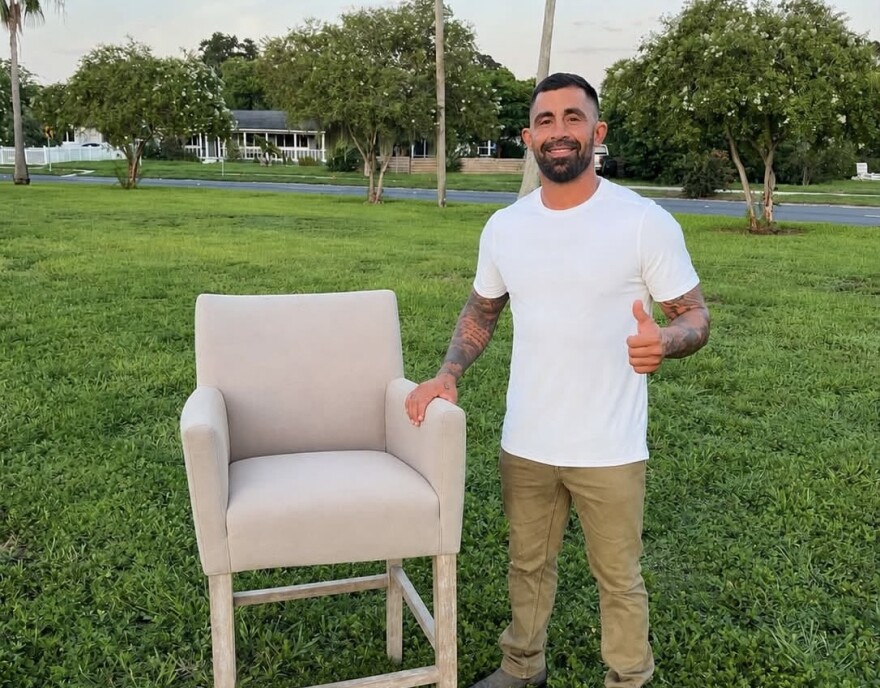 Man in white t-shirt stands outside next to a chair 