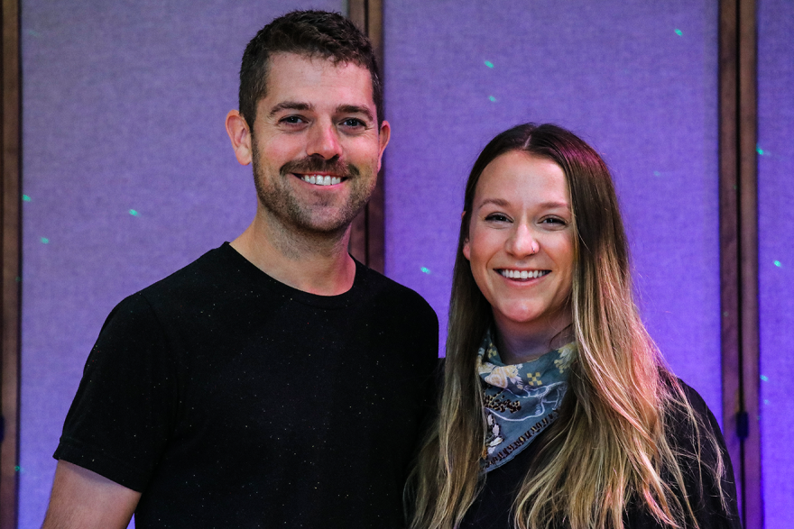 A man and woman dressed in black smile in front of a purple wall with laser dots.