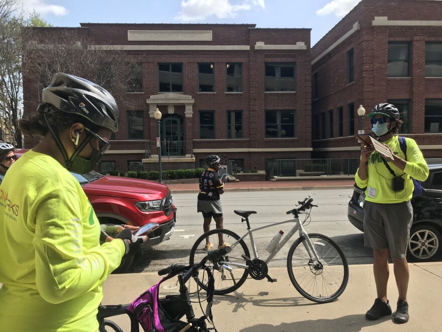 Two tour guides stand beside bicycles in bright yellow shirts. They stand in front of an old, red brick building, with a stone sign near the top that says "City-County Hospital."