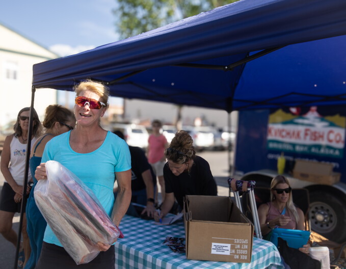 A blonde middle-aged woman in a bright blue shirt holds up a plastic bag of over foot-long salmon pieces. You can see the skin and reddish flesh. Behind here is a blue tent and blue trailer, where the company is delivering fish to other customers.