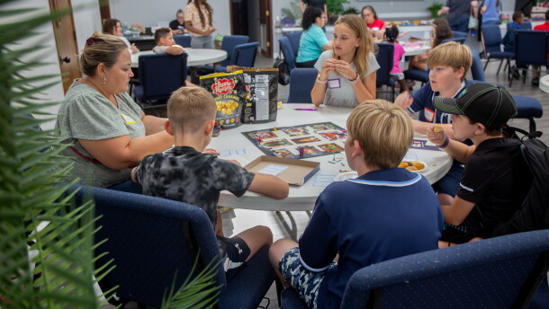 A group of five kids is sitting at a circular folding table with an adult eating chips and popcorn. They're playing the classic board game Clue.