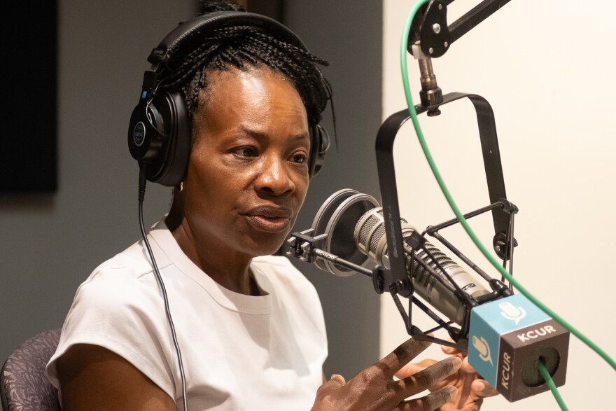 A woman inside a radio studio sits behind a microphone talking. She is gesturing with both hands.