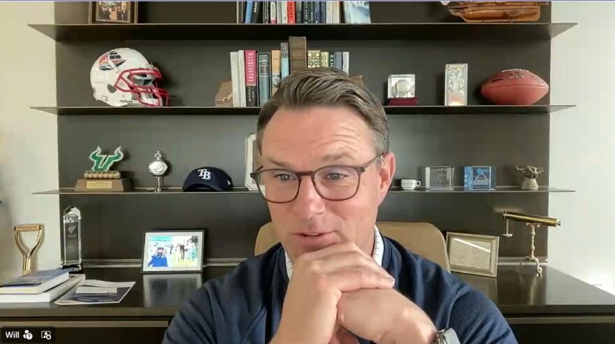 A man in glasses and navy sweater clasps hands, sitting in front of a bookcase with sports memorabilia and a dozen or so books