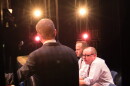 The host of an event looks at two men sitting by a table on stage, with lights shining behind them