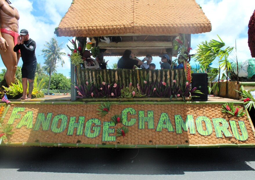 A float with a thatched roof and floral decorations displays the words “Fanohge CHamoru” during Guam’s 81st Liberation Day parade. People sit inside the float, and a man in black stands at the front, calling out to the crowd.