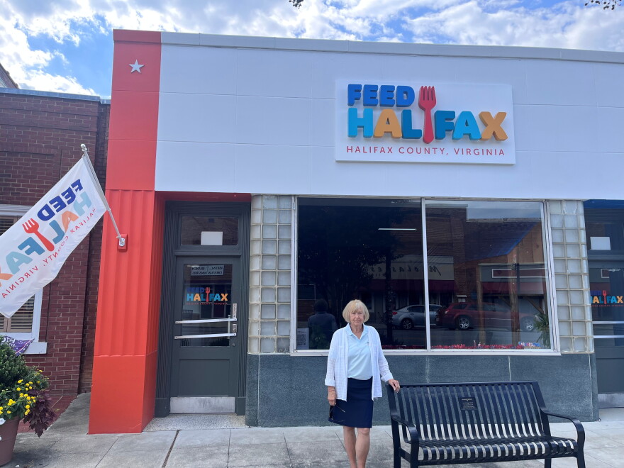 A woman wearing a blue skirt and white cardigan stands in front of a building painted in bright orange on the side and the words "Feed Halifax" on the face of the building. 