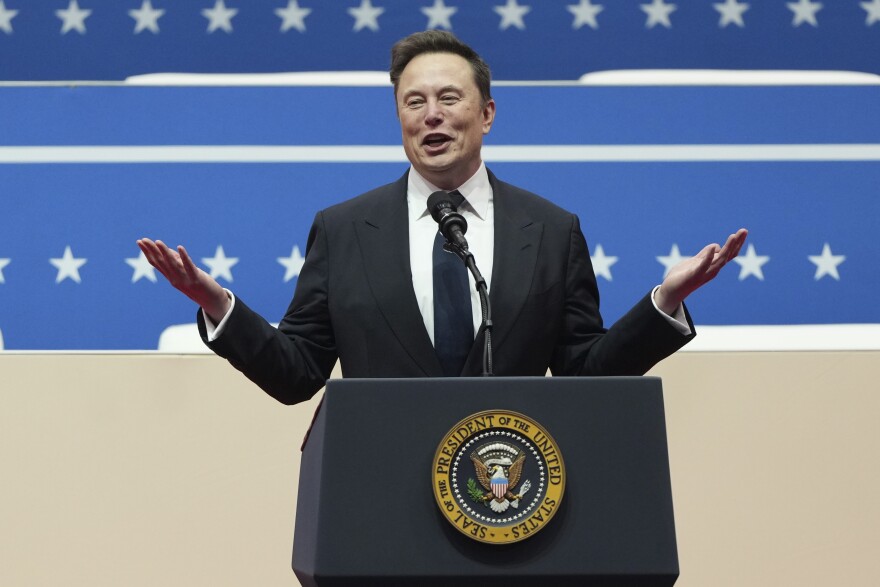 A man in a suit stands behind a podium with the U.S. presidential seal on it. Behind him is blue stripes and stars.