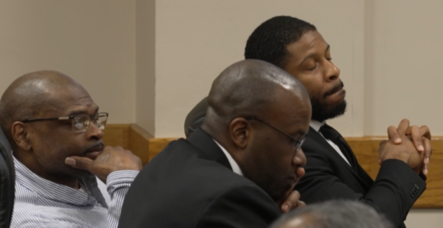 Three men sit in a courtroom, listening during proceedings.