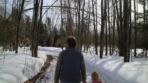 Potato farmer, Amanda Wolters holds her daughter's hand as they walk around their farm and look at damaged trees. Above them is a tree damaged so badly that it's arched over the walkway. The trails are snowy and the sky is blue. 
