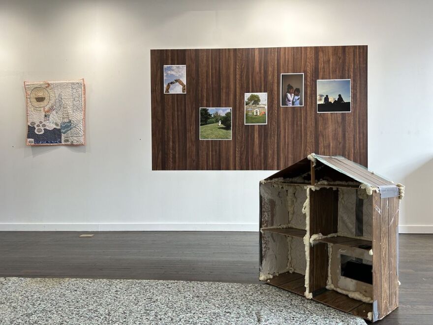 On a white wall, a quilted art work hands next to a wood panel with images on it. A dollhouse shaped sculpture of rough found material is in the foreground.