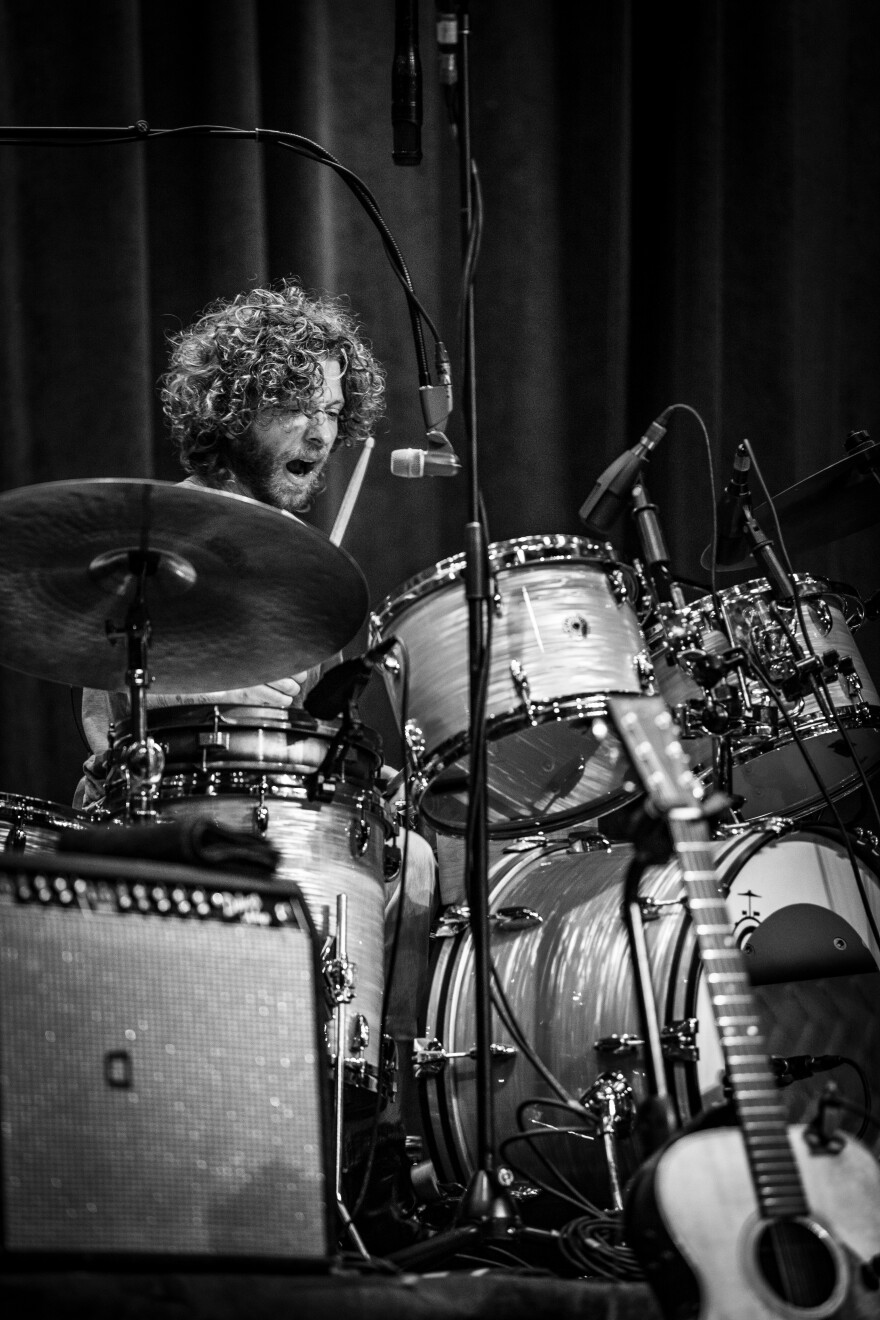 Griffin Goldsmith of the band Dawes is shown in black and white singing into a microphone while playing a drumset.