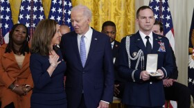 Biden and Benson are turned toward each other, smiling and talking. To their right a man stands with solemn expression, holding an open box with a medal or medals in it