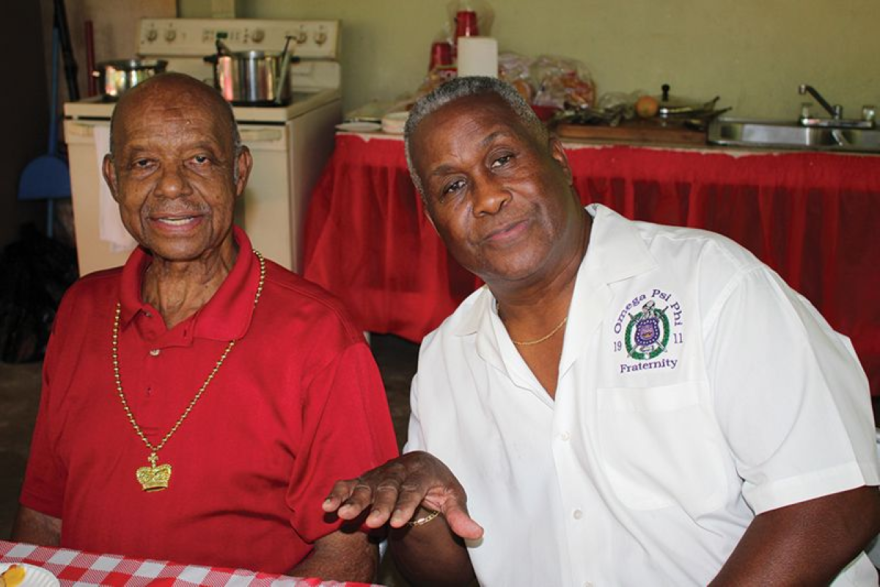 Two older Black men sitting side by side - left, man in a red polo shirt and gold necklace with a crown, and a man with gray hair and white shirt