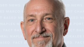 A portrait photograph of a mature, Caucasian man with a bald head and a neatly trimmed grey goatee and mustache, looking directly at the camera with a gentle, slight smile. He is wearing a dark grey, button-down shirt with a subtle, textured pattern, and a black and white paisley tie. His blue-green eyes are prominent and engaged. The background is a soft, light blue-grey color with the words "MARCO ISLAND NEWS," "NAPLES," and "CAPE CORAL" faintly repeating in a pattern. The lighting is soft and even.