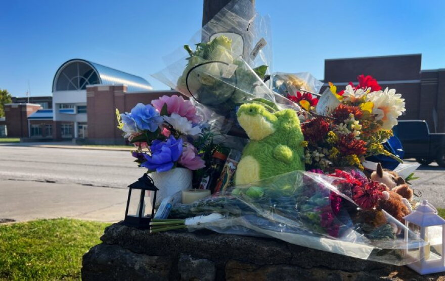 Outdoor photo shows a pile of flowers and stuff animals collecting on top of a concrete pillar. Behind, across the street, is a school building.