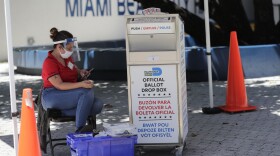 A poll worker wears personal protective equipment as she monitors a ballot drop box for mail-in ballots outside of a polling station during early voting, Friday, Aug. 7, 2020, in Miami Beach, Fla. Florida's primary election is August 18. 