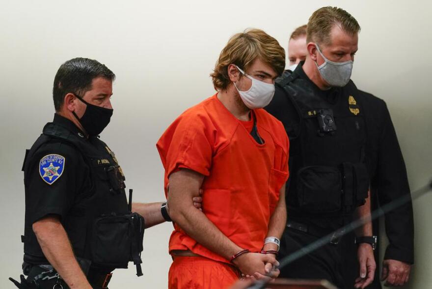 Peyton Gendron, wearing orange clothing and a white face mask, escorted by two uniformed law enforcement officers, also wearing face masks.