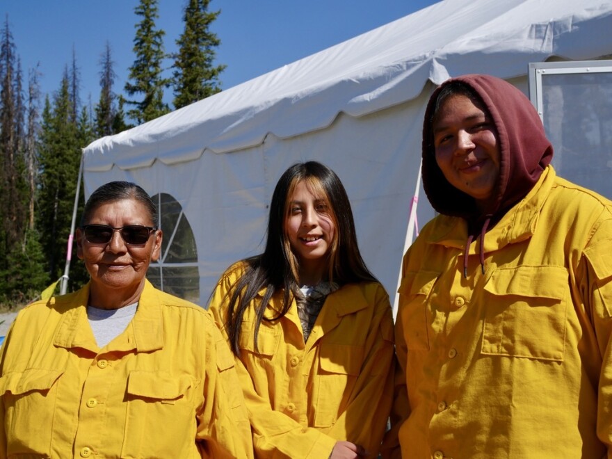 An older woman wearing dark glasses stands next to two young people in front of a large white tent, with green pine trees in the background. They are all wearing yellow button-up shirts.