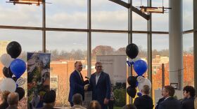 Two men in business attire shake hands at a podium during a formal announcement event, with IBM and University of Dayton signage visible, blue and white balloons decorating the space, and large windows showing autumn trees and campus buildings in the background.