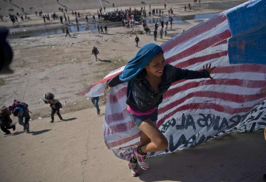 A migrant woman helps carry a handmade U.S. flag up the riverbank at the Mexico-U.S. border after getting past Mexican police at the Chaparral border crossing in Tijuana, Mexico, Sunday, Nov. 25, 2018, as a group of migrants tries to reach the U.S.