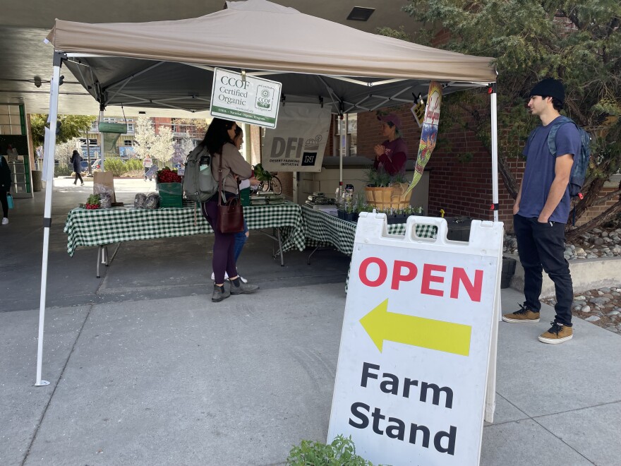 Tables with food on them are set up outside under a beige tent. There's a sign in front that says "Open" and "Farm Stand" with a yellow arrow pointing to the tented area. A couple of people are walking standing under or near the tent.