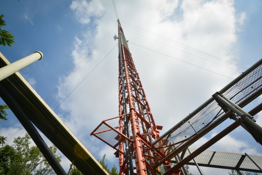 A tall, skinny steel radio tower, painted red and white is seen from below against a cloudy, blue sky. Metal framework near the base is in the foreground.