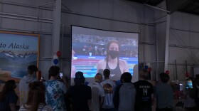 A group of people stand in a big room watching a screen with a swimmer's image projected on it.