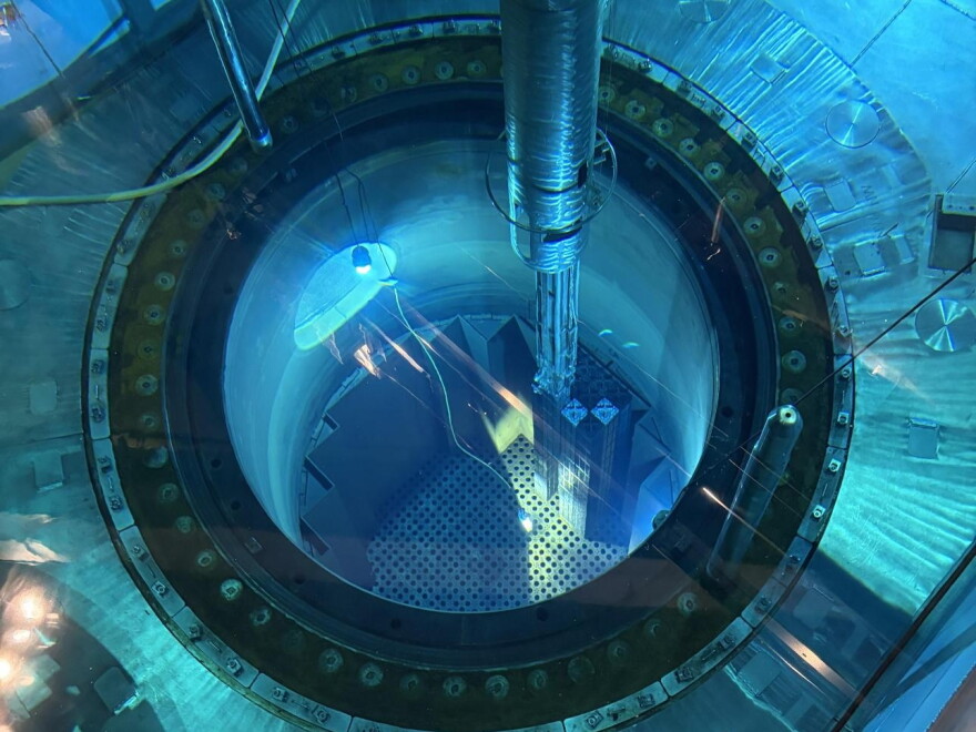 Looking down into a pool of water lit by a blue light. A metal rod extends from the top of the frame into the pool, and down into a deeper, circular pit.