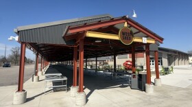 A large metal structure can be seen that looks like a long roof covering a sidewalk. It is painted red and says "west shed". Underneath it, folding tables are lined up.