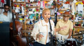 A black woman with a short blonde fade sings into a microphone while someone plays the upright base and drums behind her in front of a bookshelf.