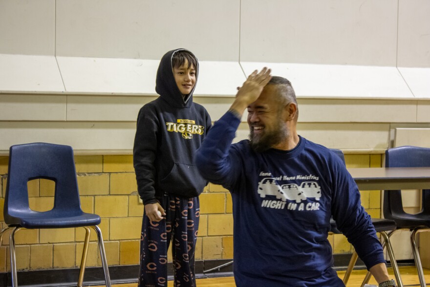 A man kneels and gestures to his forehead, where he expects his son to hit him with an egg shortly. The young boy is behind him, a ready egg in his hand.