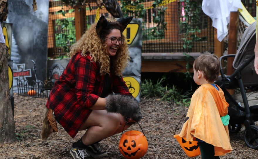 A student dressed as a werewolf hands out candy to a toddler trick-or-treater in the Monster Mash zone at the Santa Fe College Teaching Zoo's Boo at the Zoo event while laughing about their matching baskets on Wednesday, Oct. 28, 2026. (David Caine/WUFT News)