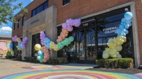 The outside of a building is decorated with garlands of balloons in various sizes and colors. The ground is painted with concentric rainbow circles. The windows read, "1160, Stonewall Columbus." 