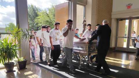 Men wearing white shirts carry a white casket into a church. People wearing pink shirts are behind them. 