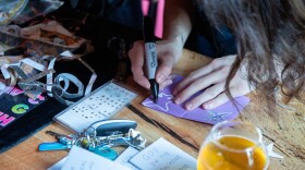 A man with long brown hair leans down close to a wooden table inside a brewery to write in Sharpie marker across a small zine made from purple construction paper. The table is littered with art supplies.
