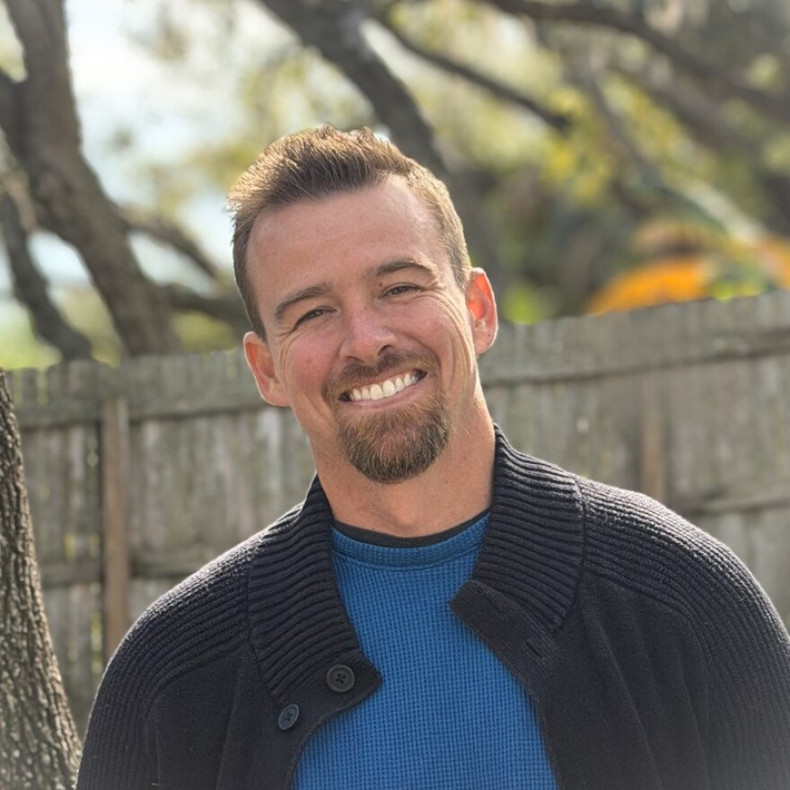 Young man standing outside in front of a brown fence wearing a blue top and black sweater