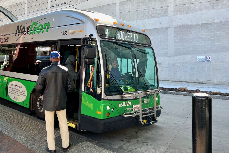 A man wearing a black coat and khaki pants stands near the door of a green and silver bus.