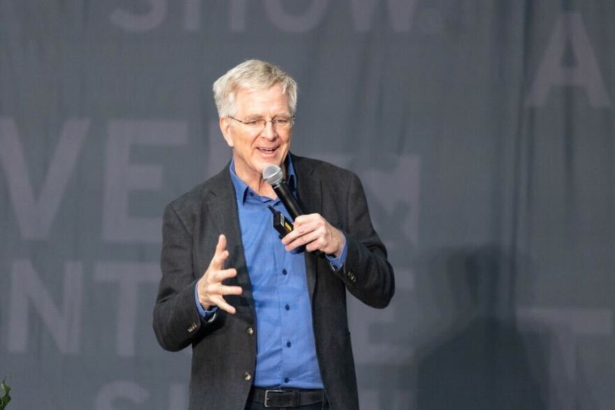 A man wearing a dark sports jacket and blue button up shirt talks into a microphone on stage.
