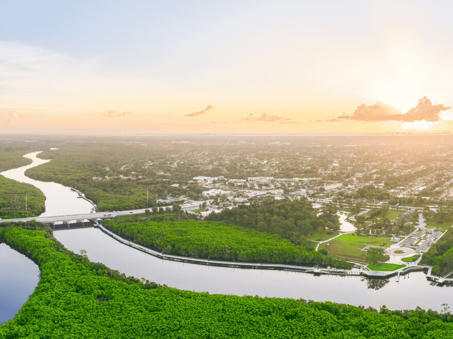 Aerial view at sunrise or sunset of a river winding through dense green mangroves, with a bridge crossing the water and a long boardwalk along the shoreline. A park with lawns, walking paths and a parking lot sits at right, with neighborhoods stretching toward a hazy horizon.