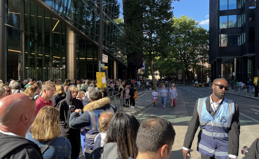 Crowds line up alongside a road, as a man in a safety vest stands over the barriers keeping them on the sidewalk.