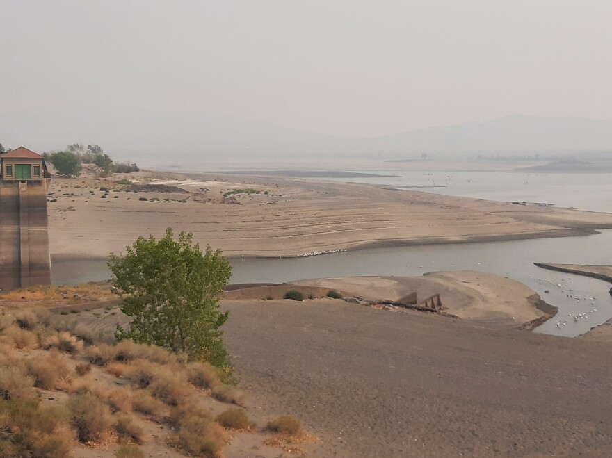 A house-like structure on concrete stilts is propped out of a very small amount of water. There are many rings on the structure and a nearby sandy beach, which shows how high the water has been in the past. There is smoke in the air from California fires.