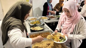 A woman is serving a dish of rice, chicken and lamb to another women who is holding a plate. There are trays of food between them. The women on the left is wearing a green headscarf and the women on the right is wearing a pink headscarf.