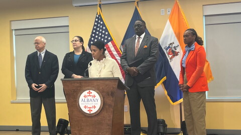 Mayor Dorcey Applyrs is standing behind a lectern with the city seal. She's flanked by two people in dress clothes on each side. 