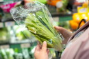 Fresh lettuce in plastic packaging held by shopper in grocery store, surrounded by various vegetables.  
