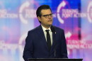 A man in a dark blue suit with white shirt and black tie stands at a lecturn or podium. He's wearing glasses and speaking into a mic.