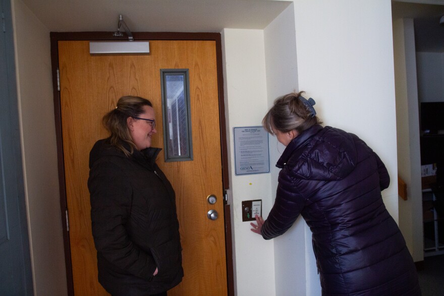 A photo showing two people inside a room standing by a wooden door. Both people are light-skinned and have greyish-blonde hair, and are wearing winter jackets. One person is pointing to a button, and the other person is looking on.