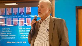 Man wearing a suit, holding a microphone before a projection screen.