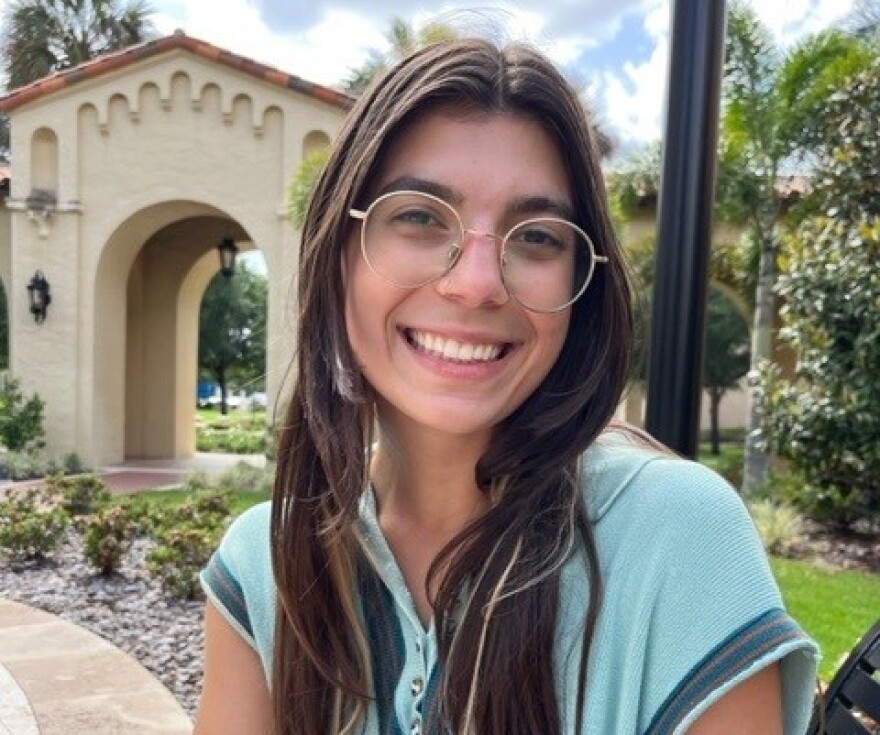 A young woman wearing glasses smiles for a picture. She's outside, with a archway and trees behind her.