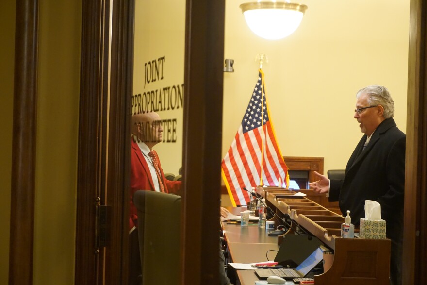 Two men talking, seen through a half open door with the words "Joint Appropriations Committee" on it.